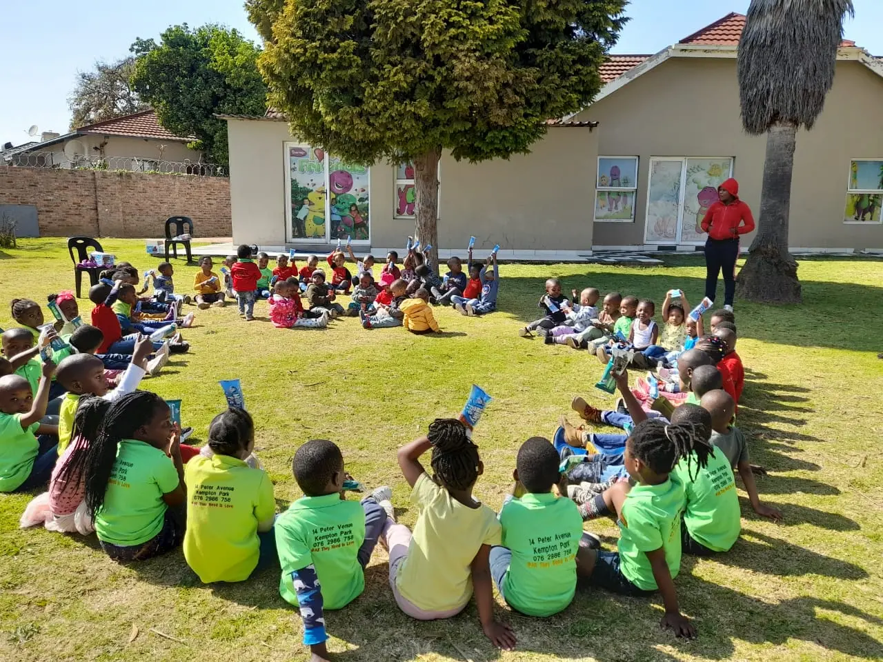 Creche learners seated down in a circle in the garden, with their teacher standing up